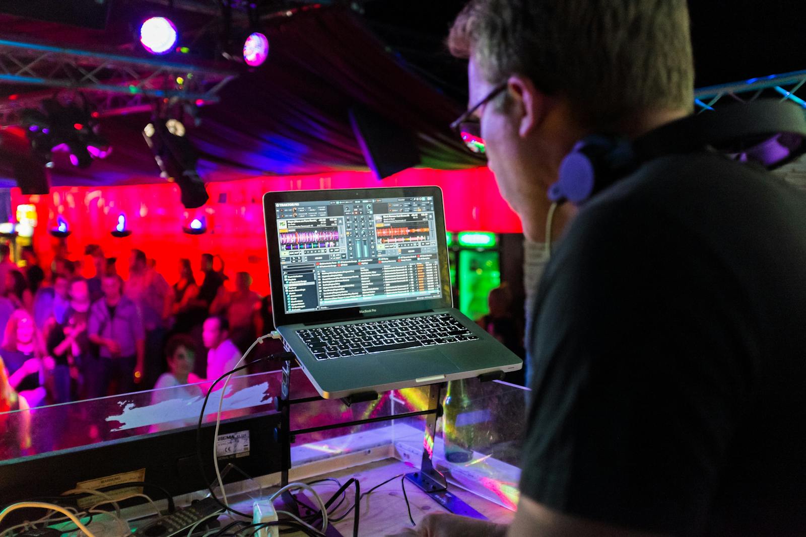 A DJ plays music using a laptop in a lively nightclub with a vibrant crowd and colorful lighting.
